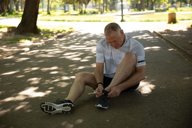 Senior man wearing sports clothes sitting on a park path tying his shoelaces. Ideal for promoting healthy lifestyles, retirement activities, fitness routines, and outdoor exercise. Useful for articles on senior fitness, health and wellness, and active aging.