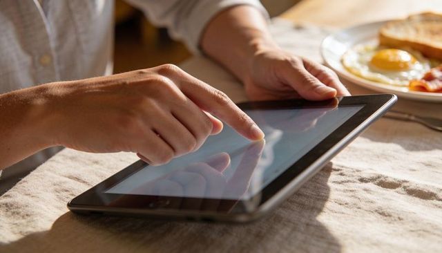 Adult Using Tablet at Breakfast Tapping Touchscreen with Eggs and Toast on Linen Table