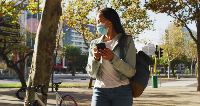 Young woman wearing a face mask is checking her phone while standing near a bicycle in an urban park. The setting is lush with trees displaying fall foliage. This image could be used for topics related to health safety, urban living, technology use in public places, and the ongoing pandemic.