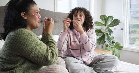 Multicultural mother and daughter applying under-eye patches while relaxing in cozy bed