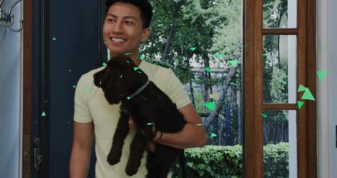 Smiling Asian man holding brown puppy at home entrance with blue door and garden view