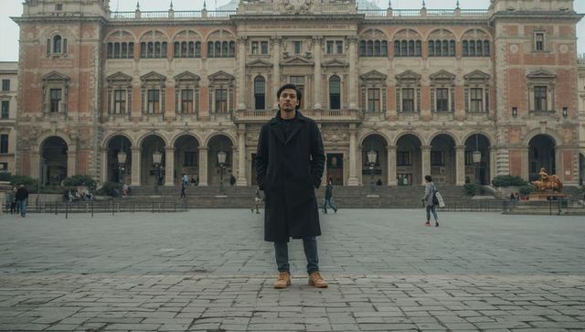 Man standing in long dark coat on historic urban plaza with colonnade and fountain