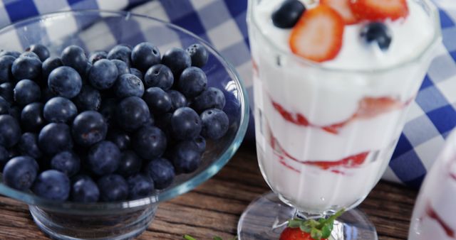 A bowl of fresh blueberries sits next to a glass of yogurt parfait layered with strawberries and blueberries, with copy space. The vibrant colors and presentation suggest a focus on healthy eating or a recipe concept.