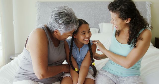 Three women from different generations, including a grandmother, mother, and daughter, sitting closely on a bed and laughing together. This can be used to illustrate family bonds, the joy of spending time with loved ones, and cross-generational relationships. Ideal for content related to family, wellness, relationships, and happiness.