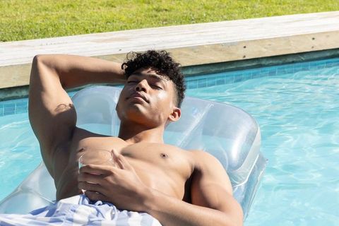 African american man lounging on clear raft in backyard pool holding drink