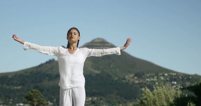 Woman stands outdoors with arms open, practicing yoga or meditation in a serene environment with a mountain in the background. Perfect for promoting health and wellness, yoga retreats, mindfulness practices, and serene nature settings.