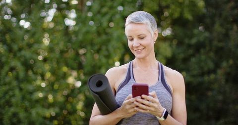 Mature woman holding yoga mat and checking smartphone while smiling in leafy park