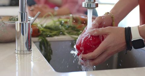 Hands rinsing fresh tomato under running kitchen faucet with smartwatch closeup, water splash