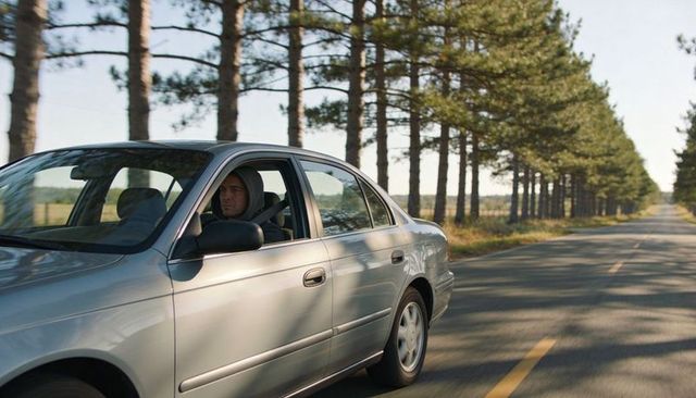 Hooded driver cruising silver sedan along sunlit pine-lined rural road with centerline