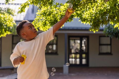African american boy blowing bubbles under sunlit tree in courtyard