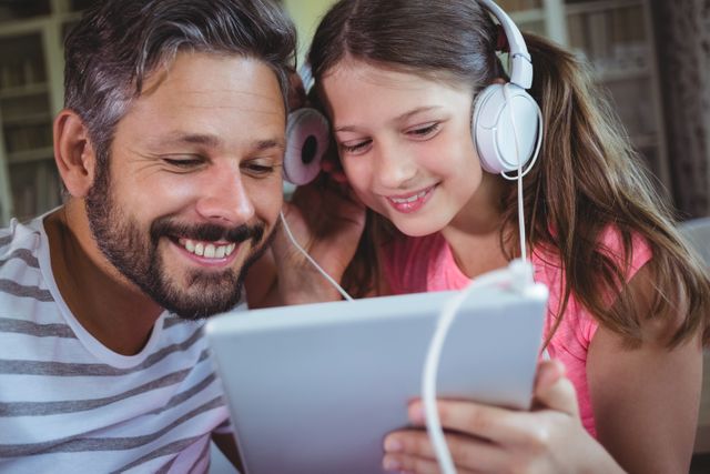 Father and daughter enjoying music together on a digital tablet at home. Both are wearing headphones and smiling, indicating a moment of bonding and relaxation. Ideal for use in family-oriented content, technology and lifestyle blogs, or advertisements promoting digital entertainment and family activities.