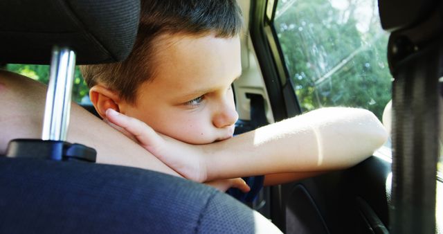 A young boy with head resting on arm, looking out car window, sunlight shining in. Ideal for concepts related to travel, family journeys, childhood moments, daydreaming, or outdoor adventures. Could be used for family travel blogs, childhood nostalgia articles, and advertisements promoting family vacations.