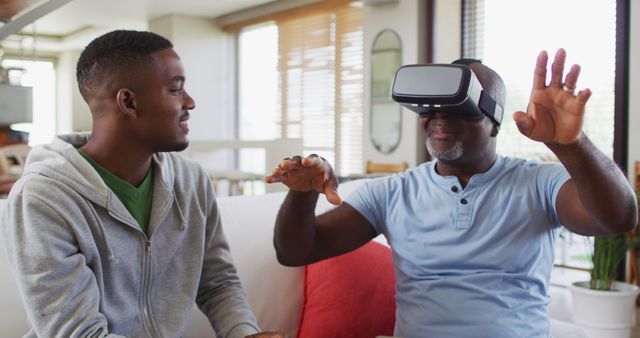 Middle-aged man sitting on sofa, wearing VR headset, experiencing virtual reality, while young adult man sits next to him, guiding and smiling. Great for depicting family interaction with modern technology, multigenerational bonding, and home leisure activities involving innovative tech.