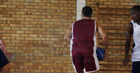 Maroon-jersey guard dribbling blue basketball past defender by padded column on indoor court