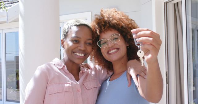 Happy diverse lesbian couple embracing holding keys outside new home. Togetherness, relationship, home ownership, moving house and domestic life, unaltered.