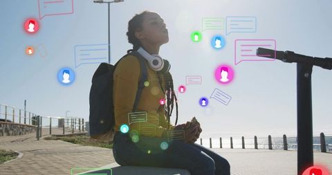 Young woman enjoying sandwich on seaside bench while surrounded by colorful chat icons