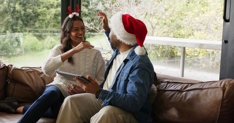 Diverse couple wearing santa hat and antlers laughing on cozy couch with tablet