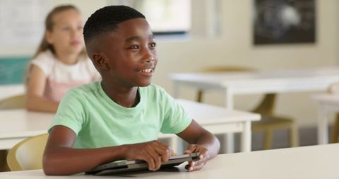 African american boy smiling and holding tablet while learning in bright modern classroom