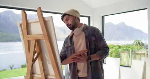 Indian artist painting at easel in bright waterside studio with mountain view
