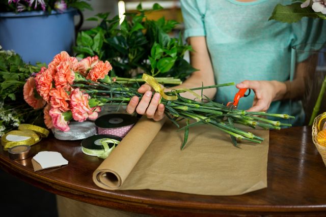 Female florist trimming flower stems while arranging a bouquet in a flower shop. Ideal for use in articles or advertisements about floristry, small businesses, creative professions, and floral design. Can also be used for content related to flower arranging tutorials, DIY projects, and the floral industry.