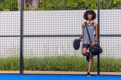 Young woman preparing for padel match on blue court, wearing headphones and carrying bag