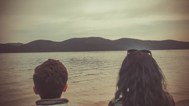 Rear view of two people, possibly friends or tourists, sitting on lakeshore admiring sunset and calm water. Great for use in travel magazines, blogs, or advertisements promoting peaceful getaways, outdoor activities, and time spent in nature or with loved ones.