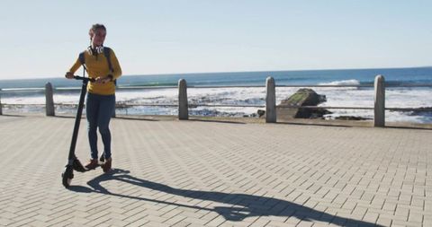 Young woman riding electric scooter along sunny seaside promenade casting long shadow