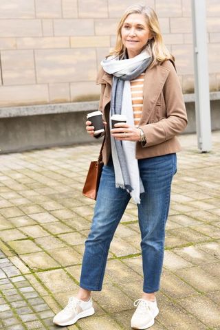 Mature woman walking in urban plaza holding two takeaway cups wearing scarf and tan jacket