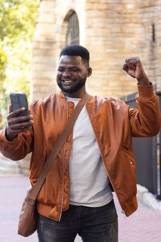African american man celebrating while checking phone, raising fist on sunlit urban walkway
