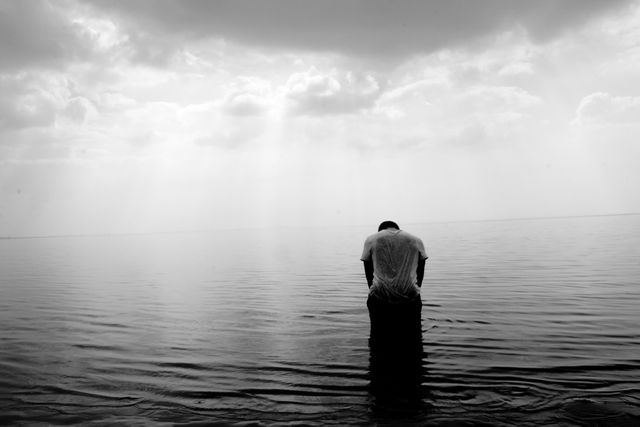 This image captures a person standing in shallow water with a dramatic sky overhead. The scene conveys solitude, reflection, and contemplation, making it suitable for themes of loneliness, peace, or personal reflection. Ideal for use in mental health, fashion editorials, inspirational posters, and artistic projects.