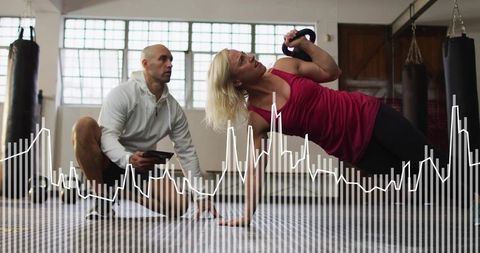 Female athlete holding kettlebell doing side plank while coach monitoring with tablet