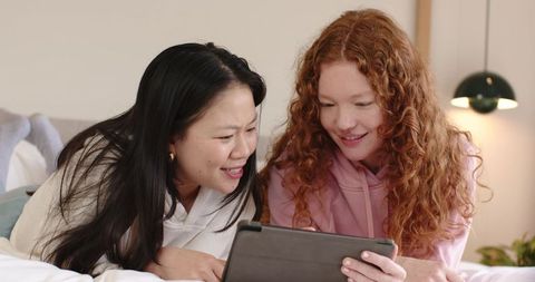 Friends sharing tablet on cozy bed laughing and browsing together at home
