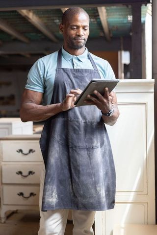 African american carpenter inspecting cabinets and using tablet in woodworking workshop