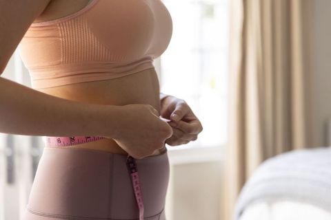 Measuring waist with pink tape wearing blush sports bra and leggings in bright bedroom