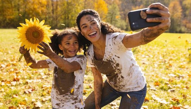 Joyful muddy mother and daughter taking sunflower selfie in autumn meadow family play