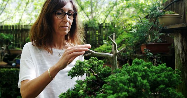 Middle-aged woman pruning a bonsai tree outdoors in a lush garden. Ideal for content related to gardening, horticulture, hobbies, nature, and environment. This image can be used in articles or advertisements focused on outdoor activities, plant care, and botany.