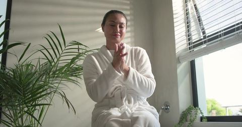 Asian woman relaxing in white bathrobe on bathtub ledge with natural light and plants