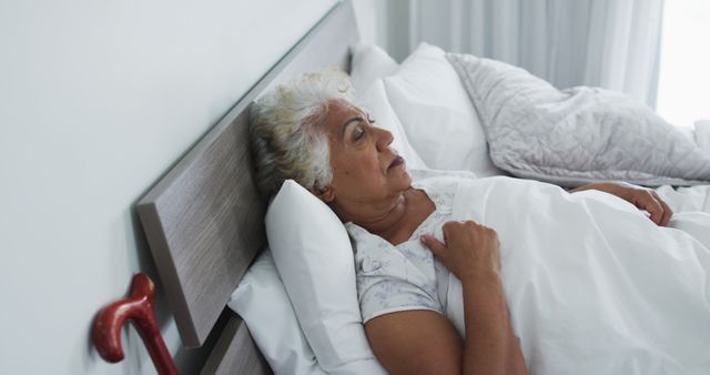 Elderly woman lies in bed covered with white blanket, with her hand on her chest and a wooden cane nearby. Ideal for healthcare, senior living, wellness, and lifestyle concepts. Suitable for illustrating retirement articles, caregiving guides, and promoting products for seniors.