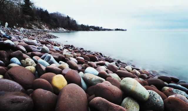 Rocky shoreline with smooth pebbles along coast under calm water and overcast sky at dusk. Peaceful coastal scene ideal for use in nature-themed projects, travel advertisements, and relaxing ambiance designs.