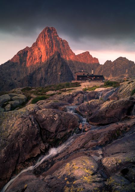 Mountain lodge illuminated by sunrise with a dramatic rocky landscape and flowing stream. Ideal for travel, adventure, and nature content. Excellent for promoting hiking destinations, scenic escapes, and outdoor retreats.