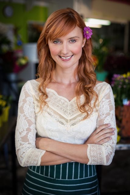 This image shows a confident female florist with red hair smiling warmly, standing in a flower shop with her arms crossed. The background is filled with various flowers and plants, creating a vibrant and welcoming atmosphere. This image is ideal for use in marketing materials for small businesses, florists, and retail, as well as in websites, blogs, and social media posts promoting customer service excellence and the beauty of fresh flowers.