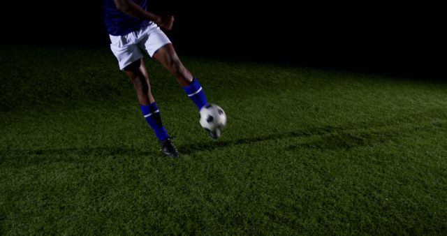 This image depicts a soccer player in blue uniform kicking the ball on a grass field at night. The focus on the player's legs and the ball highlights the action and intensity of the game. Ideal for use in sports promotions, athlete profiles, physical education resources, or advertisements related to soccer and athletic gear.