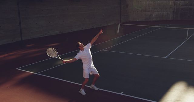 An athlete is serving during a game on an outdoor tennis court. The scene is dynamic, capturing the player in mid-action, which conveys energy and determination. This image can be used for promoting tennis events, sportswear, fitness campaigns, or sport-related content in ads, brochures, and websites.