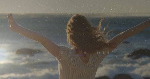 Woman stretching arms wide at rocky shoreline during golden sunlight and ocean breeze