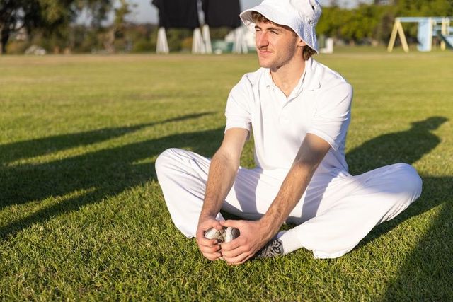 Young man sitting on grass holding metal boules in sunlit park wearing white bucket hat