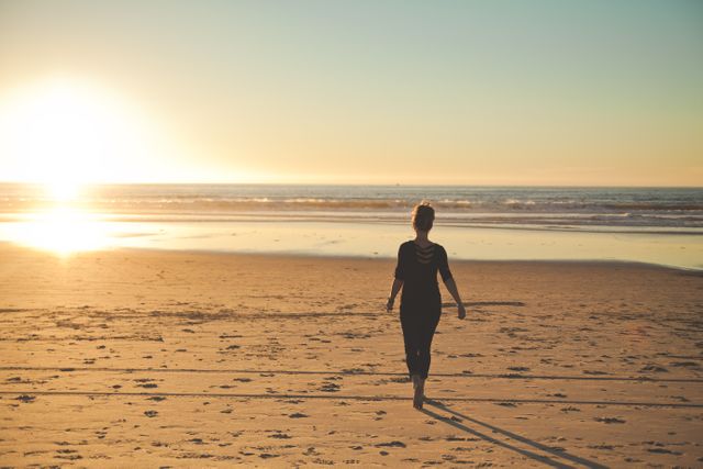 A woman walking along a sandy beach as the sun is setting over the ocean. The scene reflects serenity, calmness, and the beauty of nature, making it perfect for themes related to travel, relaxation, introspection, and vacation. Ideal for use in advertisements, travel brochures, social media posts, and websites promoting destinations.