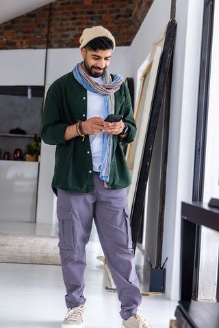 Urban artist in beanie and striped scarf using phone in bright loft studio with canvases