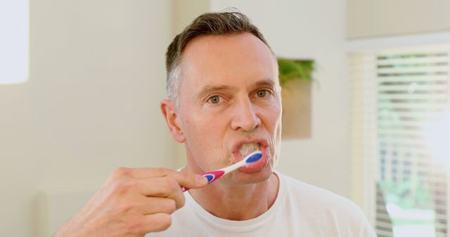 Middle-aged man brushing teeth for morning hygiene routine in a bright, home bathroom. Emphasizes dental care and healthy habits. Suitable for articles, advertisements, or publications on oral hygiene, personal care, healthy living, and dental health products.