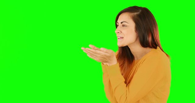 Young woman in a yellow sweater blowing a kiss to the side, standing against a green screen. Expressing affection and warmth, perfect for use in advertisements, social media content, and promotional materials requiring a friendly and approachable vibe. The green screen background allows for easy replacement with any desired backdrop.