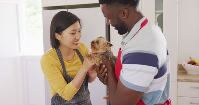 Happy diverse couple wearing aprons and petting dog in kitchen. Spending quality time at home concept.